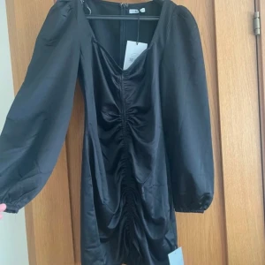 Svart kort klänning med puffärm - Supersnygg svart kort klänning från Nelly med puffiga långa ärmar och rynkade detaljer framtill. Klänningen har v-ringning och en lätt glansig finish som ger en lyxig känsla. Perfekt för dig som vill sticka ut med stil.