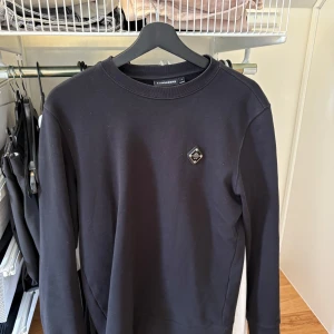 Svart sweatshirt från J.Lindeberg - Svart sweatshirt från J.Lindeberg med rund halsringning och diskret logomärke på bröstet. Tröjan har en klassisk passform och är tillverkad i mjuk bomull, perfekt för en clean och stilren look.