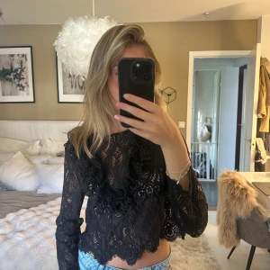 Svart spetsblus med volangdetaljer - Supersnygg svart blus i spets med långa ärmar och volangdetaljer framtill. Croppad modell med vackert mönstrad spets och rund halsringning. Perfekt att styla med jeans för en trendig look.