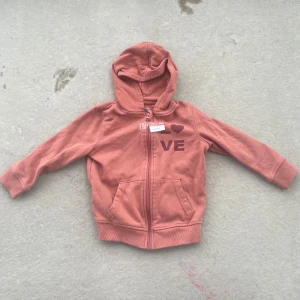 Rosa hoodie med LOVE-tryck och huva - Mysig hoodie i dämpad rosa från Juicy med dragkedja, huva och kängurufickor. Framsidan har ett tryck med texten LOVE där O:et är ett hjärta. Perfekt för chill dagar och enkel att matcha med jeans eller leggings.