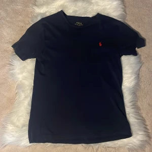 Mörkblå t-shirt från Polo Ralph Lauren - Snygg mörkblå t-shirt från Polo Ralph Lauren med klassisk röd broderad logga på bröstet. T-shirten har rund halsringning och korta ärmar. Tillverkad i mjuk bomull som känns skön mot huden. Passar om du är runt 150 