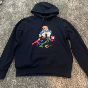 Mörkblå hoodie Polo Ralph Lauren - Säljer en mörkblå hoodie från Polo Ralph Lauren med ikoniska björnmotivet på bröstet och färgglad Polo-text. Tröjan har huva med snörning och långa ärmar. Perfekt för dig som vill ha en snygg och bekväm streetstyle-look. Nypris- 2500kr/ mitt pris- 1500kr