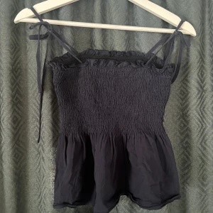 Svart smockad linnetopp - Svart linnetopp från SHEIN med smockad överdel och volangkant nertill. Toppen har tunna axelband med knyt och en lätt rynkad finish upptill. Perfekt för en trendig och luftig look under varma dagar.