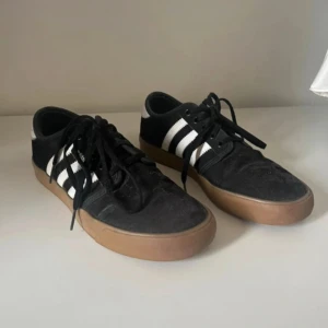 Svarta Adidas sneakers med vita ränder - Svarta Adidas sneakers med klassiska vita ränder på sidorna och vit detalj vid hälen. Skorna har brun gummisula och snörning framtill. Lite söndriga på sidan därav priset