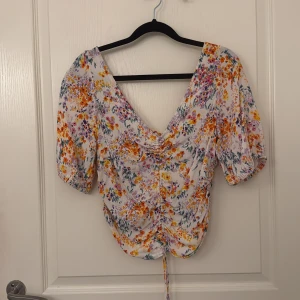 Blommig omlottopp med puffärm - Superfin blommig topp med v-ringning och puffiga korta ärmar. Toppen har omlottkänsla och snörning framtill som ger en snygg rynkad effekt. Perfekt för vår och sommar!