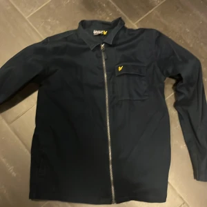 Mörkblå overshirt från Lyle & Scott - Snygg mörkblå overshirt från Lyle & Scott med klassisk krage, bröstficka med gul logga och hel dragkedja framtill. 