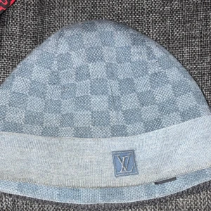 Grå  Louis Vuitton mössa - Snygg grå mössa från Louis Vuitton med klassiskt rutigt mönster och LV-logga framtill. Mössan är stickad och har en bred uppvikt kant för extra stilpoäng. Perfekt för dig som vill ha en exklusiv look även när det är kallt ute.
