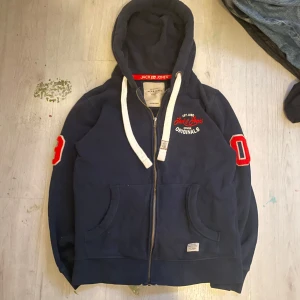 Mörkblå hoodie från Jack & Jones - Mörkblå hoodie från Jack & Jones med vit snörning och dragkedja. Röd broderad logga på bröstet och röda detaljer på ärmarna. Mjuk bomullskänsla, huva och praktiska fickor framtill. Perfekt för chill dagar och streetstyle.