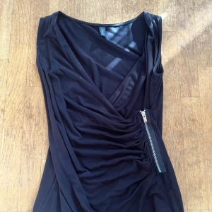 Svart omlottopp från Kaul Edition - Svart omlottopp från Kaul Edition i storlek S. Toppen har en djup v-ringning, är ärmlös och har en snygg dragkedjedetalj på sidan. Materialet är mjukt och stretchigt, perfekt för en trendig och stilren look.