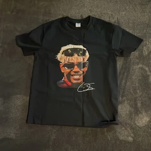 Svart t-shirt med tryck och signatur - Svart t-shirt från Gildan med coolt tryck på bröstet som visar en person med blont hår, solglasögon och bandana. Under motivet finns en vit signatur. Klassisk passform och rund hals. Perfekt för dig som gillar statement-plagg.