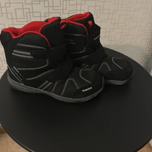 Svarta vinterkängor från Treksta - Svarta kängor från Treksta med grå och vita detaljer samt röd insida. Snygg sportig design med kardborreband och robust sula. Perfekta för kalla dagar och outdoor-äventyr. Materialet är syntet och textil, vilket gör dem både lätta och slitstarka.