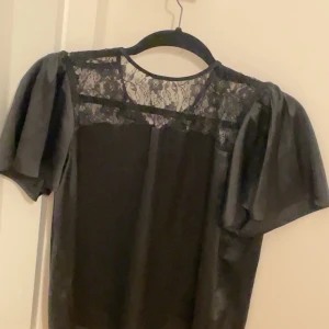 Svart blus med spets från JDY - Säljer en svart blus från JDY i storlek M. Toppen har korta puffärmar och en transparent överdel i blommig spets. Materialet är mjukt och lätt, perfekt för att lyfta din outfit med en elegant touch.