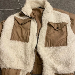Beige teddyjacka från Boohoo - Säljer en beige teddyjacka från Boohoo med vita fluffiga paneler och två stora fickor framtill. Jackan har dragkedja, hög krage och långa ärmar i beige polyester. Perfekt för dig som vill ha en mysig och trendig look under kyliga dagar.