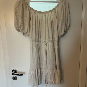 Beige kortklänning från WEEKDAY - Superfin beige kortklänning från WEEKDAY med puffiga korta ärmar, offshoulder-modell och volang nertill. Klänningen har knyt i midjan och är gjord i ett mjukt bomullsliknande material. Perfekt för sommarens alla tillfällen!