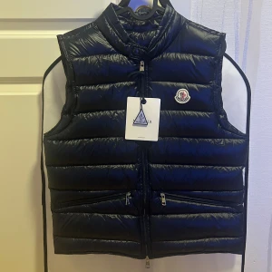 Moncler Väst - Snygg mörkblå dunväst från Moncler med klassisk quiltad design, hög krage och dragkedja framtill. Västen har två fickor med dragkedja och ikonisk Moncler-logga på bröstet. Perfekt för lager-på-lager och stilren look.