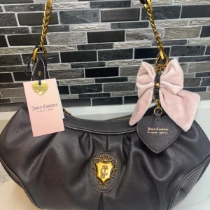 Juicy Couture axelväska med rosett - Mörkbrun axelväska från Juicy Couture i skinn med guldfärgade detaljer och kedja. Väskan har en stor ljusrosa rosett och hjärtformad tag med logga. Invändigt foder med Juicy Couture-tryck. Perfekt för dig som gillar statement-accessoarer.