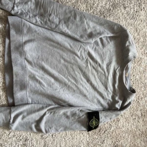 Stone Island tröja - Säljer en grå sweatshirt från Stone Island med klassisk rund hals och ribbade muddar. Tröjan har den ikoniska svarta logotypen med gul brodyr på vänster ärm. Perfekt basic med snygg streetkänsla.
