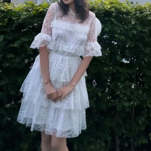 Vit spetsklänning med volanger - Superfin vit kortklänning i spets med volanger och hög hals. Klänningen har trekvartsärm med volangdetaljer och flera lager i kjolen som ger en romantisk och luftig känsla. Perfekt för sommarens alla tillfällen! perfekt som studentklänning