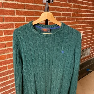 Grön kabelstickad tröja från Morris - Mörkgrön kabelstickad tröja från Morris med rund halsringning och diskret blå logga broderad på bröstet. Tröjan är långärmad och gjord i mjuk bomull, perfekt för lager på lager under kyliga dagar.