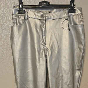 Silvriga byxor i skinnimitation L/XL - Snygga byxor i metallic silver med klassisk femficksmodell och bälteshällor. Byxorna har en rak passform och är tillverkade i skinnimitation som ger en cool och trendig look. Perfekta för dig som vill sticka ut med en futuristisk vibe.