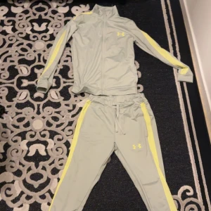 Under Armour träningsjacka ljusgrå S - Säljer en ljusgrå träningsjacka från Under Armour med gula detaljer längs ärmarna och dragkedja framtill. Jackan har lång ärm, hög krage och är tillverkad i mjukt syntetmaterial. Perfekt för träning eller sportiga outfits. Inga skador osv!!!! Sällsynt!!!!