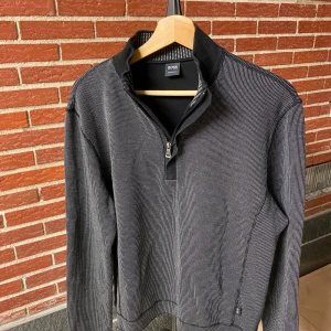 Grå half zip tröja från Hugo Boss - Snygg grå långärmad tröja från Hugo Boss med diskret mönster och svart krage. Tröjan har half zip-dragkedja och förstärkta svarta partier på armbågarna. Perfekt för dig som gillar stilrena och klassiska plagg med en modern touch.