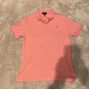 Rosa pikétröja från Ralph Lauren - Snygg rosa pikétröja från Polo Ralph Lauren med klassisk krage och två knappar framtill. Ikoniska broderade loggan på bröstet. Skön och mjuk bomull, perfekt till jeans eller shorts. Enkel och stilren design som alltid funkar.