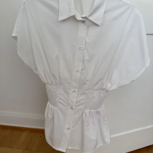 Vit skjorta med smock från H&M - Snygg vit skjorta från H&M med korta ärmar och smockade partier i midjan.