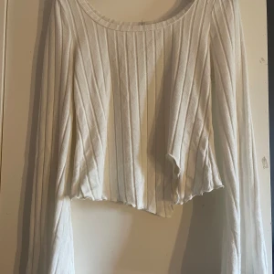 Vit ribbad croppad långärmad topp - Säljer en vit långärmad topp med ribbat mönster och croppad passform. Toppen har utsvängda ärmar och en vågig, asymmetrisk nederkant som ger en cool och trendig look. Perfekt för dig som gillar stilrena men ändå unika plagg.