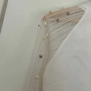 Vit blus med pärldetaljer - Säljer en vit blus med transparenta, långärmade ärmar i mesh med tunna ränder och pärlor i olika storlekar längs axlar och ärmar. Blusen har hög hals och en stilren, modern look som sticker ut med sina unika detaljer.