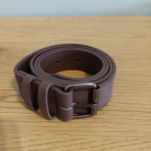 Brunt skärp i skinn med metallspänne - Säljer ett stilrent brunt bälte i skinn med klassiskt silverfärgat metallspänne. Bältet har en enkel design utan mönster och passar perfekt till jeans eller chinos. Bältet är rakt och har flera hål för justering.
