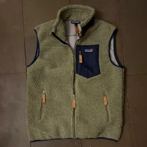 Patagonia fleeceväst - Mysig olivgrön fleeceväst från Patagonia med hög krage, tre fickor med dragkedja och detaljer i mörkblått tyg. Västen har en bröstficka med logga och snygga läderdetaljer på dragkedjorna. Perfekt lager-på-lager-plagg för kyliga dagar. Knappt använd och därmed väldigt bra skick. Storlek M men sitter mer som S 