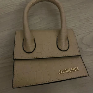 Beige  handväska i skinn - Superfin beige handväska med krokopräglat mönster och guldfärgad logga framtill. Väskan har en stilren, boxig form och två rejäla handtag. Perfekt accessoar för att lyfta din outfit.