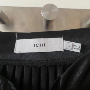 Svart plisserad kjol från ICHI - Snygg svart plisserad kjol från ICHI i glansigt material. Kjolen har ett klassiskt midilångt snitt och breda plisseringar som ger ett coolt fall. Perfekt att styla med chunky sneakers eller boots för en trendig look.