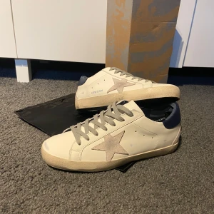 Golden Goose  - Snygga vita sneakers från Golden Goose superstar med grå mockadetaljer och ikonisk stjärna på sidan. Skorna är i storlek 43 och allt medföljer som finns på bild. Skorna är helt nya aldrig använda, hör av er vid frågor 😁Detta är den mest eftertraktade modellen ny pris 6000kr