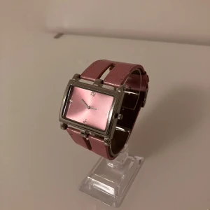 Rektangulär rosa klocka med skinnband - Stilren klocka med rektangulärt rosa ur och matchande rosa skinnarmband. Urtavlan har minimalistiska siffror och silverfärgade visare. Klockan har en bred rem med vita sömmar och metallinfattning runt urtavlan. Perfekt accessoar för dig som gillar färg och unika former. Behöver ett nytt batteri.