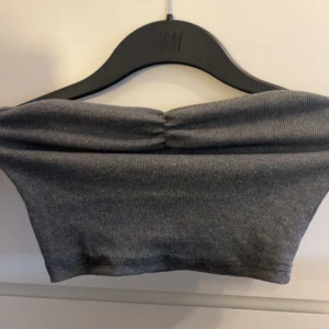Grå ribbad bandeau tubtopp H&M - Säljer en grå bandeau tubtopp från H&M. Toppen är ribbad och har en snygg rynkning framtill som ger en cool detalj. Perfekt att styla med höga jeans eller kjol för en trendig look.
