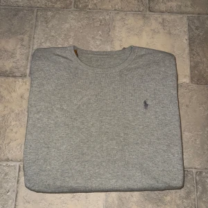 Grå stickad tröja från Ralph Lauren - Snygg grå stickad tröja från Ralph Lauren med klassisk rund halsringning och broderad logga på bröstet. Perfekt basic med lång ärm och ribbade muddar, passar till allt och är superenkel att styla.