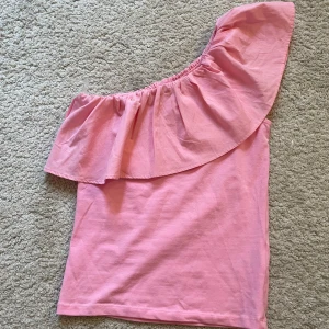 Rosa volang topp från Kappahl - Snygg rosa oneshoulder topp från Kappahl med bred volang över axeln. Toppen är ärmlös på ena sidan och har en tight passform. Använd en gång. Storlek 146/152. Skriv privat för frågor 💕buda gärna!