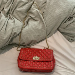 Röd quiltad axelväska med nitar - Snygg röd axelväska i quiltat skinn med guldiga nitar och kedjerem. Väskan har ett klassiskt lock med vridlås och insidan är rymlig nog för det viktigaste. Perfekt för dig som vill sticka ut med en edgy och trendig accessoar.