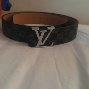 Bälte  - Säljer ett bälte med klassiskt rutigt mönster i svart och grått. Bältet har en bred form och en silverfärgad metallspänne med en logga. Insidan är brun och materialet är skinn. Perfekt för dig som vill ha en lyxig och stilren accessoar. Det kommer med box och påse