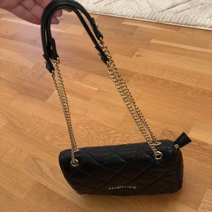 Svart quiltad axelväska från Valentino - Snygg svart axelväska från Valentino med quiltat mönster och guldfärgade kedjor som axelrem. Väskan är tillverkad i skinnimitation och har ett stilrent, lyxigt uttryck med logga framtill. Perfekt storlek för det viktigaste och en riktig statement piece. Köpt på zalando . Öppen för prisförslag men ej för lågt för den är i för bra skick. 