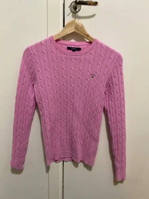 Rosa kabelstickad tröja från Gant - Säljer en rosa kabelstickad tröja från Gant med rund halsringning och broderad logga på bröstet. Tröjan har långa ärmar och ribbade muddar. Perfekt för dig som gillar klassisk och stilren design.