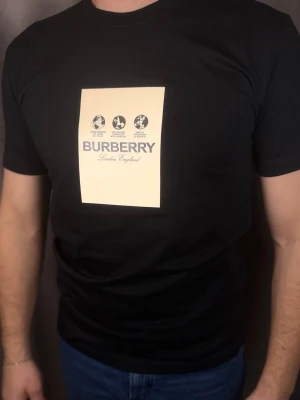 Svart Burberry t-shirt med tryck - Svart t-shirt från Burberry med ett stort beige tryck på bröstet där det står 'Burberry London England' och grafiska symboler ovanför. T-shirten har rund hals och korta ärmar. Perfekt för dig som gillar stilrena och exklusiva plagg.