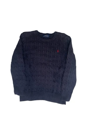 Mörkblå kabelstickad tröja Polo - Kabelstickad mörkblå tröja från Polo Ralph Lauren med rund halsringning och röd broderad logga på bröstet. Klassisk modell med ribbade muddar och nederkant. Perfekt för en stilren look.