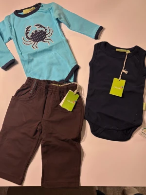 Klädpaket pojke 62/68 - Säljer ett set med en långärmad blå body med krabbtryck, en ärmlös marinblå body och ett par bruna byxor. Alla plagg har mjukt material och bekväm passform. 