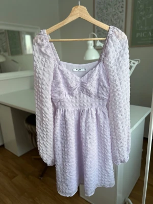 Ljuslila kort klänning från NA-KD - Supersöt ljuslila kort klänning från NA-KD med puffiga långärmar och strukturerat tyg. Klänningen har en markerad midja, v-ringning och en söt rosettdetalj framtill. Perfekt för dig som vill ha en feminin och trendig look.