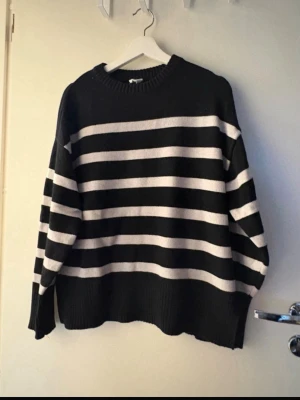 Svartvit randig stickad tröja - Säljer en oversized stickad tröja med breda svarta och vita horisontella ränder. Tröjan har rund halsringning, långa ärmar och ribbade muddar samt slitsar i sidorna. Perfekt för en avslappnad och trendig look.