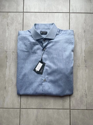 Ljusblå skjorta från Tiger of Sweden - Snygg ljusblå skjorta från Tiger of Sweden med diskret prickigt mönster. Klassisk krage, knäppning framtill och långa ärmar med manschett. Perfekt för en stilren look och tillverkad i bomull för en skön känsla. Vid frågor eller funderingar skriv gärna!