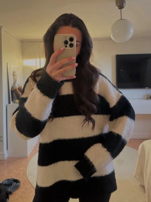 Randig svartvit stickad tröja - Mysig stickad tröja med breda svarta och vita ränder. Tröjan har en avslappnad passform och långa ärmar. Perfekt för att hålla sig varm under kyliga dagar. Materialet ser fluffigt och mjukt ut.